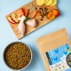 Ein Holzschneidebrett hält rohe Hähnchenkeulen, Lachs, Apfelscheiben, Aprikosen, Samen und eine Schüssel mit Haustierfutter neben einem beschrifteten Beutel.