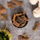 Eine schwarze Schüssel mit Hundeleckerlis, verstreute Jerky-Stücke, ein weißes Seilspielzeug und eine kleine Pflanze in einem geflochtenen Korb in der Nähe.