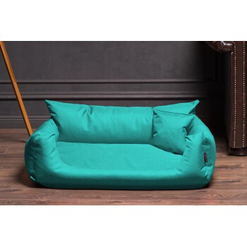 Türkisfarbenes Haustierbett mit erhöhten Seiten und zwei Kissen, auf Holzboden vor einer dunklen Wand platziert.