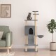 Eine getigerte Katze sitzt auf einem hohen grauen Kratzbaum mit mehreren Ebenen, einem gemütlichen Versteck und einer hängenden Schüssel, neben einem grünen Sofa und einer Topfpflanze.