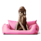 Ein brauner Hund sitzt aufrecht auf einem plüschigen rosa Hundebett und schaut neugierig zur Seite.