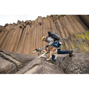 Ein Mann in einer grauen Jacke und einem gelben Helm erklimmt felsiges Terrain mit einem braun-weißen Hund, der ein blaues Geschirr trägt.