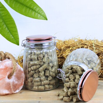 Zwei Glasbehälter mit Haustierfutterpellets, einer umgekippt und auf Holz verschüttet, mit einem rosa Salzkristall und Stroh im Hintergrund.