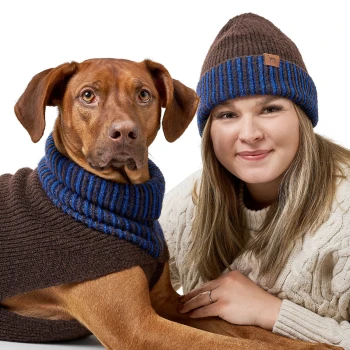 Ein Hund trägt einen braunen Pullover mit blauen Streifen und sitzt neben einer Frau in einem cremefarbenen, kabelgestrickten Pullover, beide sehen gemütlich aus.