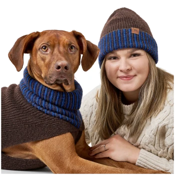 Ein Hund trägt einen braunen Pullover mit blauen Streifen und sitzt neben einer Frau in einem cremefarbenen, kabelgestrickten Pullover, beide sehen gemütlich aus.