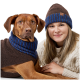 Ein Hund trägt einen braunen Pullover mit blauen Streifen und sitzt neben einer Frau in einem cremefarbenen, kabelgestrickten Pullover, beide sehen gemütlich aus.