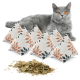 Eine graue Katze mit leuchtend orangefarbenen Augen liegt hinter fünf dekorativen Kissen, gefüllt mit Katzenminze und mit Blattmustern, und getrocknete Katzenminze ist davor verstreut.