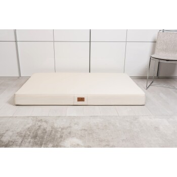 Ein cremefarbenes Haustierbett mit einem Lederetikett, auf einem hellen Holzboden neben einem grauen Teppich und einem modernen Stuhl platziert.