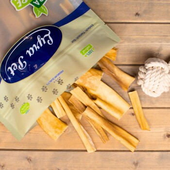 Eine Tüte mit Haustierleckerlis, beschriftet mit "100% Natürlich," die verschiedene Streifen von kaubaren Leckerlis auf eine Holzoberfläche auskippt, neben einem weißen Seilspielzeug.