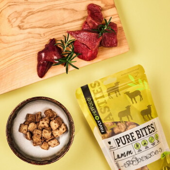 Ein brauner Hund mit schlappen Ohren schaut auf eine Tüte "Pure Bites" Lamm-Snacks, die Texte über Transparenz, einzelnes Protein und getreidefrei enthält.
