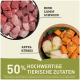 Schüssel mit rohen Fleischwürfeln (Rind, Lamm, Schwein) neben einem Teller mit Apfel und Kürbis, mit dem Text '50% hochwertige tierische Zutaten.'