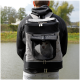Eine Person in einem schwarzen Hoodie trägt einen grauen Haustier-Rucksack mit einem Netzfenster, das einen kleinen Hund zeigt, am Ufer eines Sees.