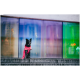 Ein schwarzer Hund sitzt in einem leuchtend pinken Regenmantel auf Steinstufen vor einem bunten, verschwommenen Glas-Hintergrund.