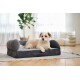 Curly cream dog (face blurred) lying on gray bolster pet bed with brown leather tag on rug in bright living room.
