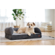 Lockiger cremefarbener Hund (Gesicht unscharf) liegt auf einem grauen Bolster-Tierbett mit braunem Lederetikett auf einem Teppich in einem hellen Wohnzimmer.