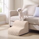 Kleiner weißer Hund steht auf einem beigefarbenen Sofa neben beige gerippten Haustierstufen in einem sonnendurchfluteten Wohnzimmer.
