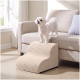 Kleiner weißer Hund steht auf einem beigefarbenen Sofa neben beige gerippten Haustierstufen in einem sonnendurchfluteten Wohnzimmer.