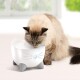 Un chat duveteux avec un pelage clair boit d'une fontaine à eau en forme de chat blanche avec des pattes grises et des motifs de moustaches.