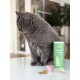 Graue Katze sitzt auf dem Tisch neben einer grünen Dose 'KatKare Shiny Cat Topping', rosa Schaufel und einem Haufen Pulver.