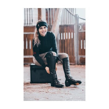 Eine Person sitzt auf einer schwarzen Kiste in einer Scheune, trägt ein schwarzes Langarmshirt und hohe Reitstiefel und passt ihr Schuhwerk an.