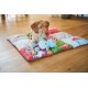 Puppy lying on a colorful quilted pet mat on a wooden floor, front paws holding a blue plush bear with a red heart.