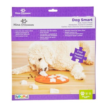 Ein flauschiger weißer Hund interagiert mit einem Puzzle-Spielzeug auf einer orangefarbenen Matte, während die Verpackung "Dog Smart" mit Anweisungen in mehreren Sprachen zeigt.
