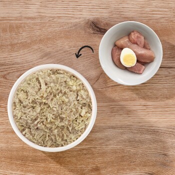 Eine Schüssel mit feuchtem Haustierfutter mit stückiger Textur steht auf einer Holzoberfläche, neben einem kleinen Teller mit rohen Hühnerstücken und einem gekochten Ei.