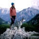 Eine Frau in einer rosa Fleecejacke und blauen Mütze steht auf einem Felsen mit einem grau-weißen Hund an der Leine und blickt auf eine bergige Landschaft.