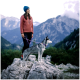 Eine Frau in einer rosa Fleecejacke und blauen Mütze steht auf einem Felsen mit einem grau-weißen Hund an der Leine und blickt auf eine bergige Landschaft.