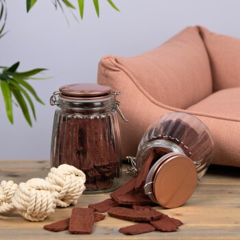 Ein Glasbehälter voller Hundeleckerlis, der auf einen Holztisch fällt, neben einem geknoteten Seilspielzeug und einem gemütlichen rosa Haustierbett im Hintergrund.