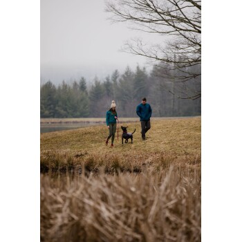 Eine Frau in einer türkisfarbenen Jacke und ein Mann in einer blauen Jacke führen einen schwarzen Hund auf einem grasbewachsenen Weg in der Nähe eines Sees, umgeben von Bäumen.