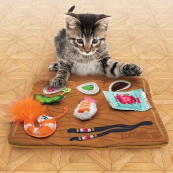 Un chaton tabby joueur interagit avec un tapis de sushi en feutre coloré comportant des articles en peluche sushi, y compris des crevettes, du poisson et des baguettes.