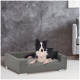 Ein schwarz-weißer Border Collie ruht sich bequem auf einem modernen grauen Hundebett mit einem kleinen schwarzen Kissen aus, vor einer strukturierten grauen Wand.