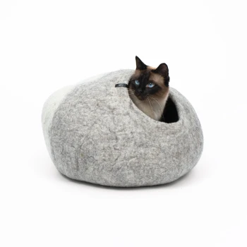 Eine Siamkatze mit blauen Augen, die aus einem gemütlichen, grauen Filz-Haustierbett in Form eines Steins schaut, vor einem weißen Hintergrund.