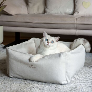 Ein gemütliches, helles Katzenbett mit weicher Textur, mit dem Logo "The Cattery" auf der Seite gestickt, mit einer Katze, die darin liegt.
