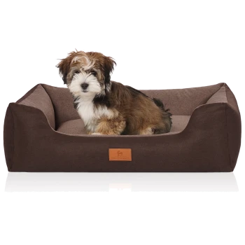 Flauschiger braun-weißer Welpe, der in einem rechteckigen braunen Hundebett mit Lederaufnäher mit der Aufschrift KNUFFELWUFF sitzt.