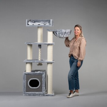 Eine Frau in einer beigen Bluse und blauen Jeans hält eine graue Hängematte neben einem hohen, mehrstöckigen Katzenbaum mit Kratzpfosten.