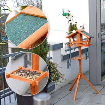 Holz-Vogelfutterspender mit grünem Dach, gefüllt mit Samen, auf einem Dreibeinständer; dekorativer Pfau-Windspinner im Hintergrund.