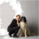 Eine Person in schwarzer Kleidung kniet neben einem großen, flauschigen Bernhardinerhund mit schwarz-weißen Fell, beide lächeln vor einem hellgrauen Hintergrund.