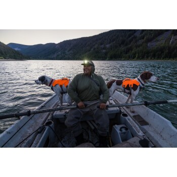 Eine Person in einem Boot mit zwei Hunden, die leuchtend orangefarbene Schwimmwesten tragen, umgeben von einem ruhigen See und bewaldeten Bergen bei Dämmerung.