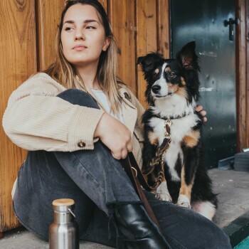 Eine Frau sitzt auf einer Holzplattform mit ihrem schwarz-braunen Hund, beide schauen in die Kamera. Eine Metallwasserflasche ist in der Nähe.