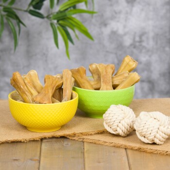 Zwei Glasbehälter gefüllt mit Hundeknochen, einer umgekippt, neben zwei Seilspielzeugen auf einer Holzoberfläche, mit einem rosa Haustierbett im Hintergrund.