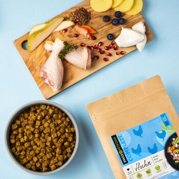 Eine Schüssel mit Haustierfutterpellets steht neben einem Holzschneidebrett mit Hähnchenschenkeln, Obst, Gemüse und Samen.