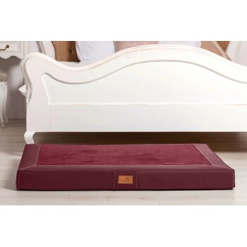 Ein burgunderfarbenes Haustierbett mit einer glatten Oberfläche und genähten Kanten, auf einem hellen Holzboden unter einem weißen Bettgestell platziert.