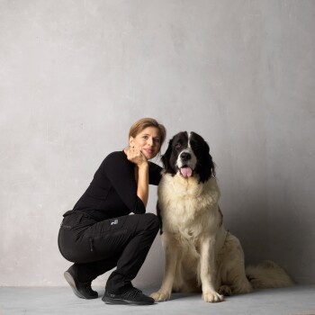 Eine Person in schwarzer Kleidung kniet neben einem großen, flauschigen Bernhardiner mit schwarz-weißem Fell, beide lächeln vor einem grauen Hintergrund.
