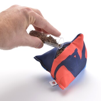 Eine Hand gießt getrocknete Kräuter aus einem Glasrohr in ein buntes Haustierspielzeug in Form eines Kissens mit einem blauen und orangefarbenen Design.