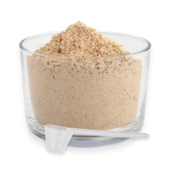 Eine Glasschüssel gefüllt mit hellbraunem Pulver, neben der eine kleine weiße Messschaufel liegt.