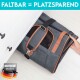 Eine graue Tragetasche für Haustiere mit braunen Lederelementen, die teilweise unter einem Schrank hervorschaut, mit "FALTBAR = PLATZSPAREND" und einem "DEUTSCHE MARKE"-Logo.
