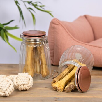 Zwei Glasbehälter gefüllt mit Hundekauartikeln, einer umgekippt, neben zwei weißen Seilspielzeugen auf einer Holzoberfläche, mit einem rosa Haustierbett im Hintergrund.