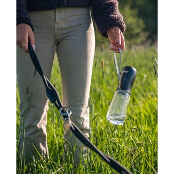 Eine Person, die in einer Hand eine Hundeleine und in der anderen eine tragbare Wasserflasche hält, steht auf einem grasbewachsenen Feld.